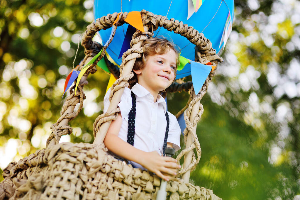 маленький хлопчик з кучерявим волоссям з телескопом в руках на тлі синього кошика повітряної кулі посміхається і дивиться на відстань
. - Фото, зображення