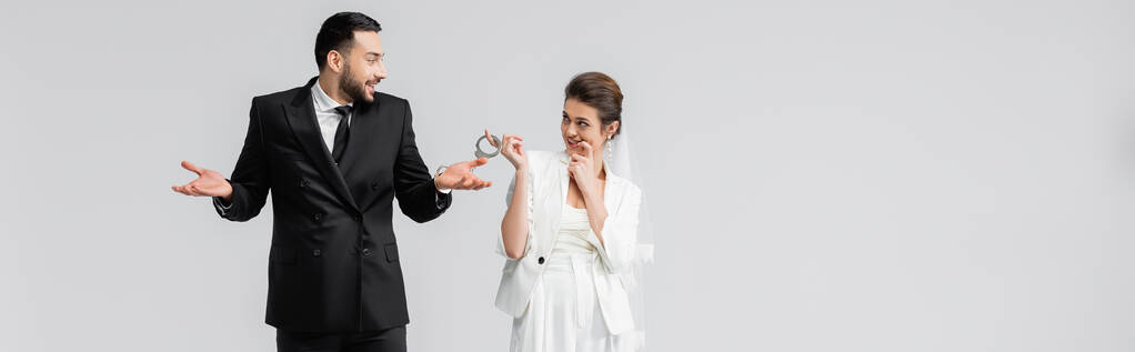 Mariée rusée regardant le marié arabe menotté montrant un geste d'haussement d'épaules isolé sur gris, bannière  - Photo, image