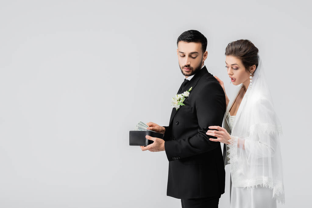 Mariée excitée debout près du marié arabe avec de l'argent et portefeuille isolé sur gris  - Photo, image