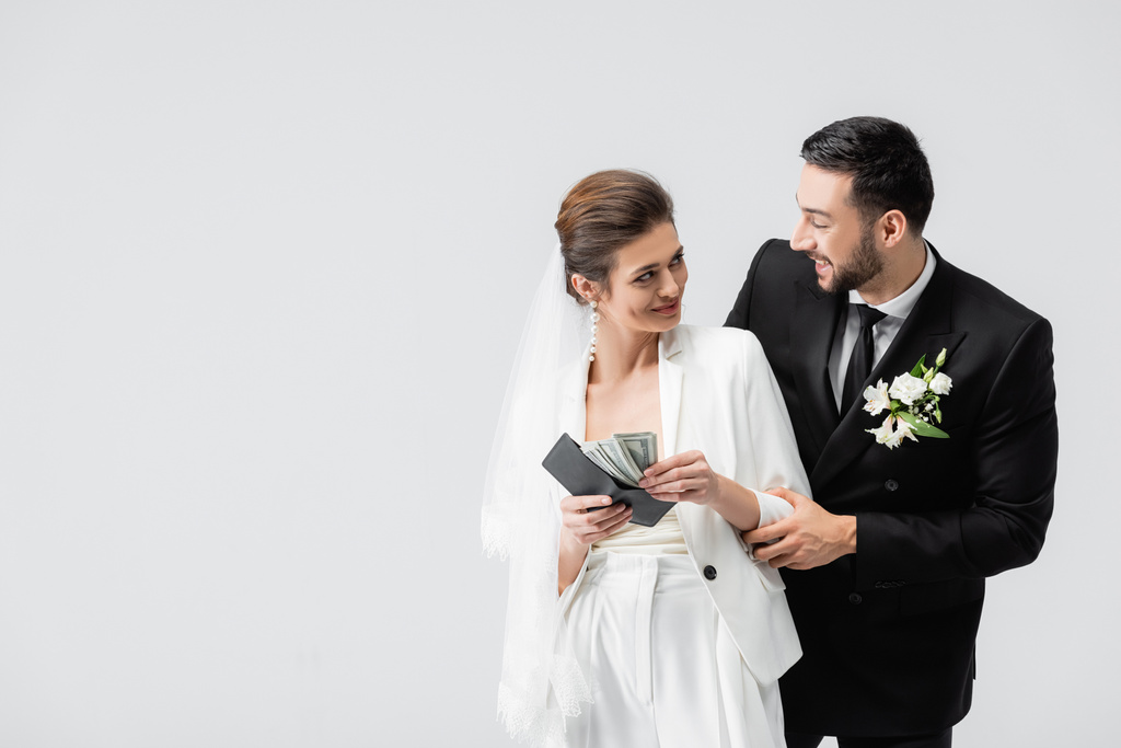 Smiling muslim groom standing near bride with dollar banknotes and wallet isolated on grey  - Photo, Image