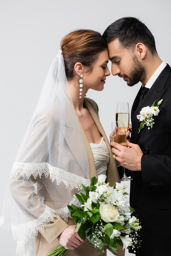 elegantní nevěsta a mladý arabský snoubenec stojí tváří v tvář se zavřenýma očima izolovaných na šedé - Fotografie, Obrázek