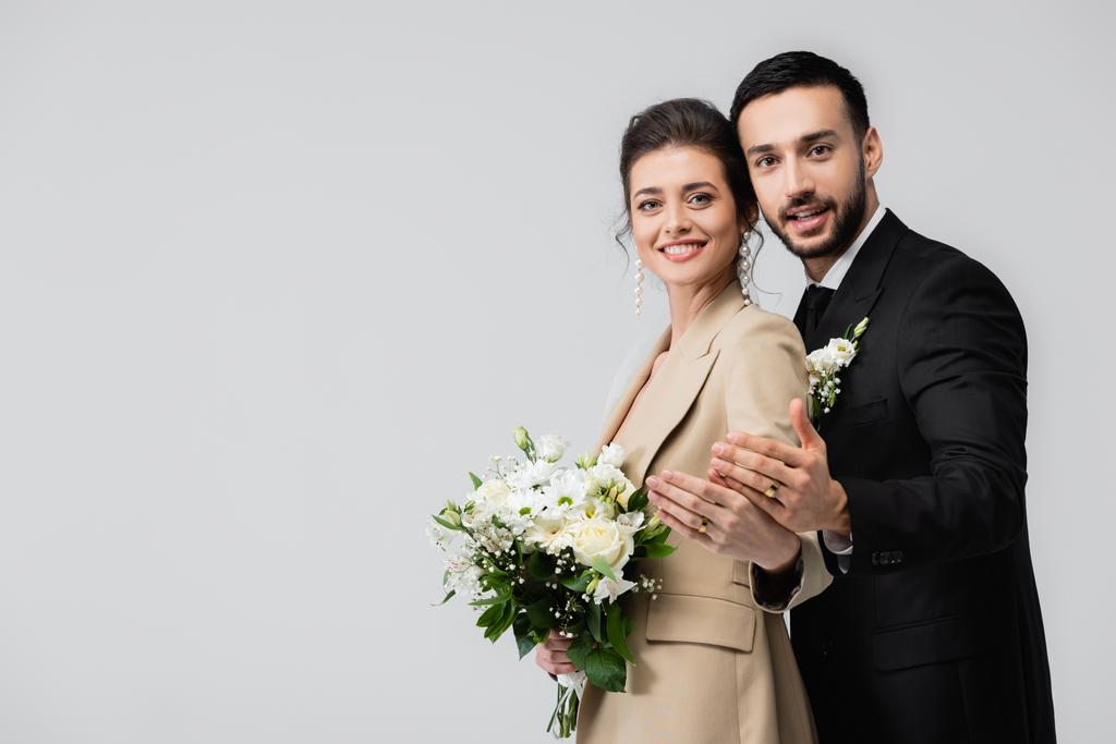 joyful multiethnic newlyweds looking at camera while showing wedding rings isolated on grey - Photo, Image