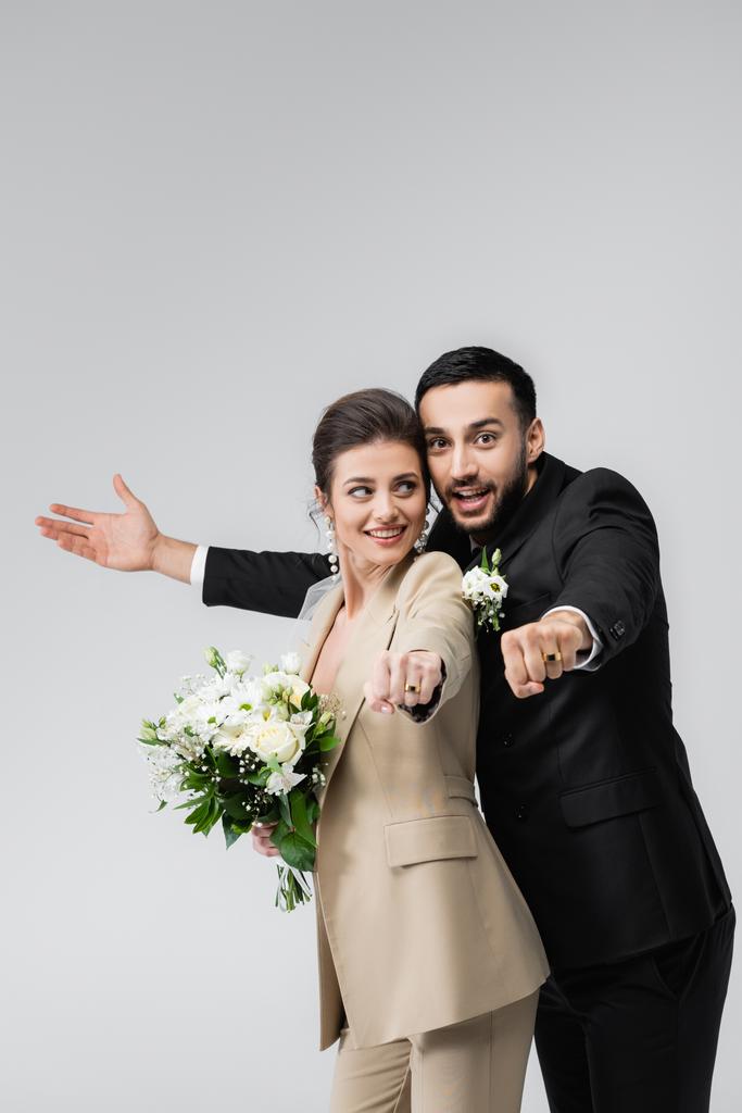 excitada, pareja recién casada multiétnica mostrando anillos de boda aislados en gris - Foto, Imagen