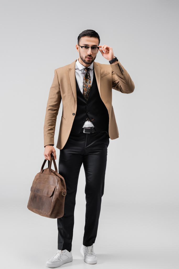 Модный мужчина в очках в полный рост позирует с кожаной сумкой на сером фоне - Фото, изображение