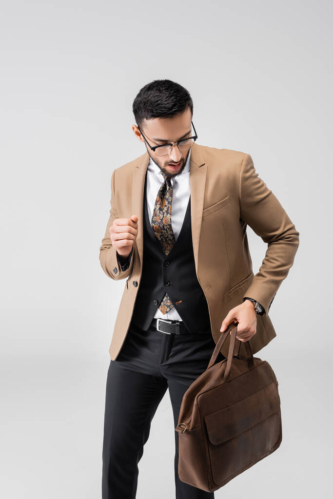 młody arabski mężczyzna w modnym garniturze spacery ze skórzaną torbą izolowane na szarości - Zdjęcie, obraz