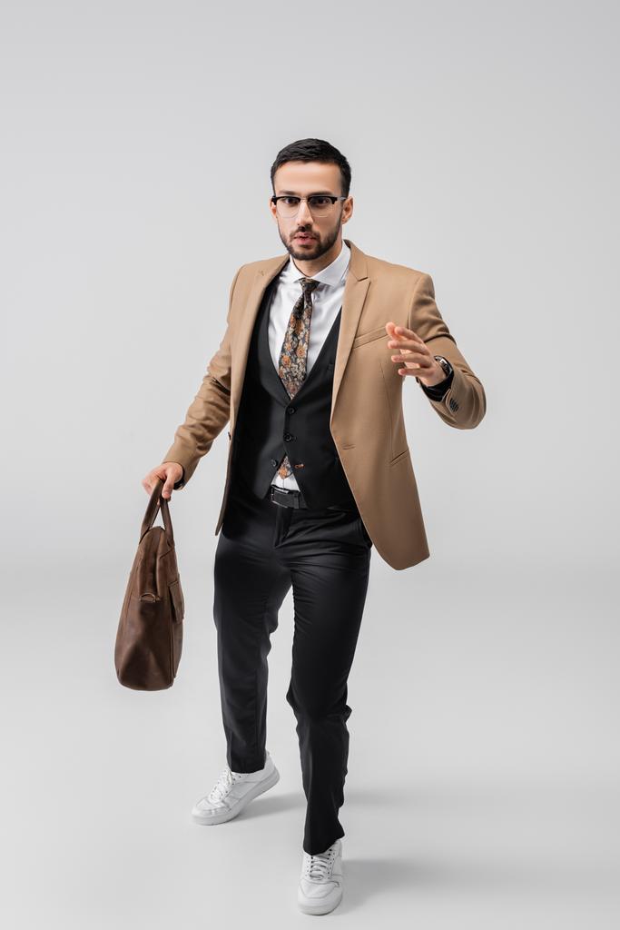 vista completa de hombre árabe con estilo mirando a la cámara mientras camina con bolsa de cuero en gris - Foto, imagen