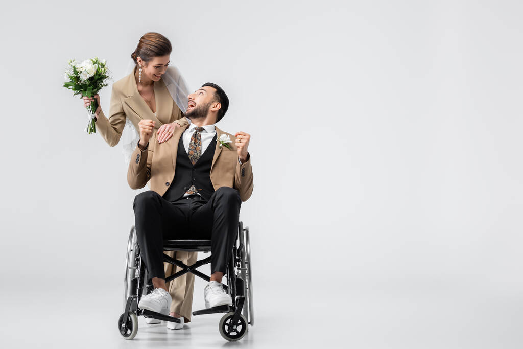 excited, handicapped arabian man showing win gesture while looking at happy bride on grey - Photo, Image