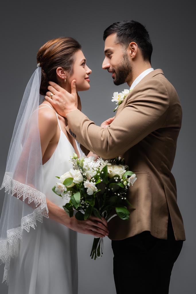 вид збоку щасливого арабського нареченого, що обіймає шию елегантної нареченої на сірому
 - Фото, зображення