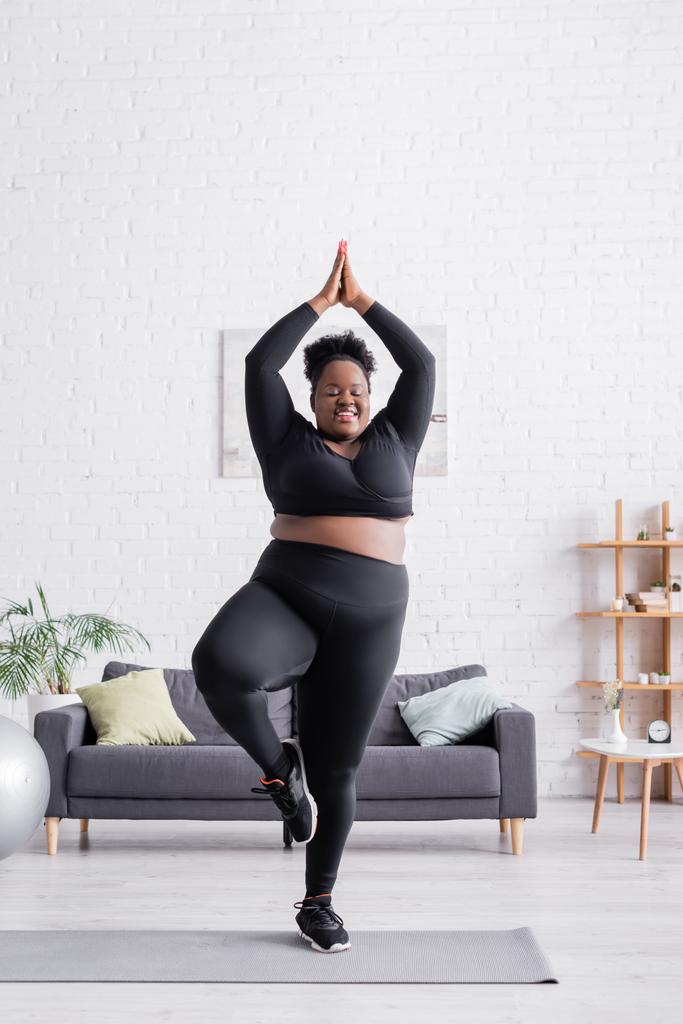 gelukkig Afrikaans amerikaans plus size vrouw in sportkleding staande in yoga pose in woonkamer - Foto, afbeelding
