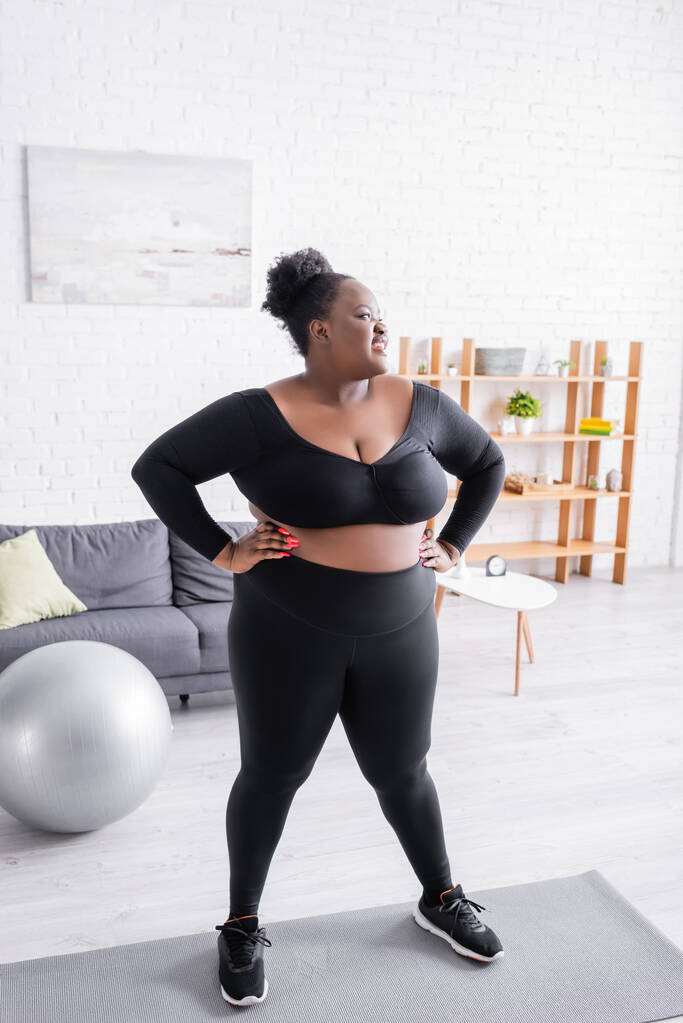 heureux afro-américain plus taille femme en vêtements de sport debout avec les mains sur les hanches dans le salon - Photo, image
