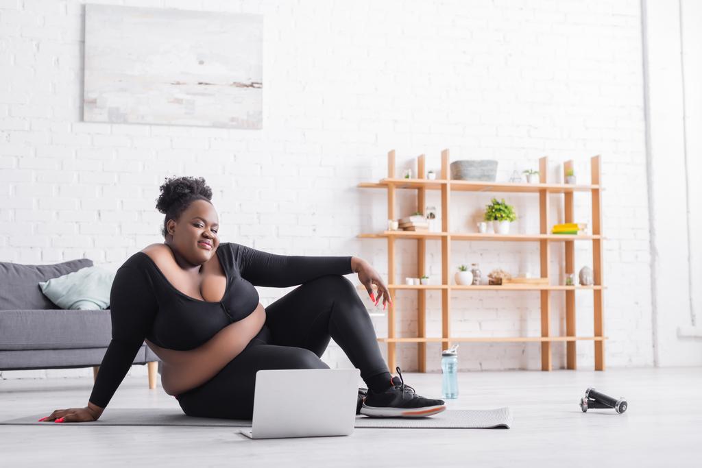hymyilevä afrikkalainen amerikkalainen plus koko nainen katsomassa online-koulutusta kannettavalla tietokoneella istuen kunto matto  - Valokuva, kuva