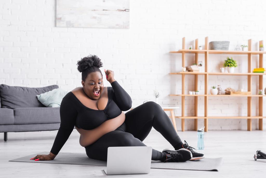 podekscytowany Afryki plus rozmiar kobieta w odzieży sportowej siedzi na macie fitness i patrząc na laptopa w salonie - Zdjęcie, obraz