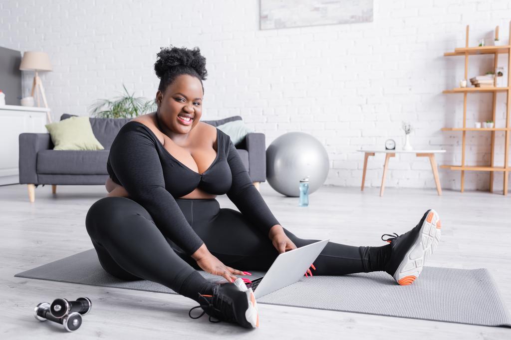 ευτυχισμένη Αφροαμερικανή συν μέγεθος γυναίκα σε αθλητικά είδη κάθεται στο στρώμα γυμναστικής με φορητό υπολογιστή  - Φωτογραφία, εικόνα