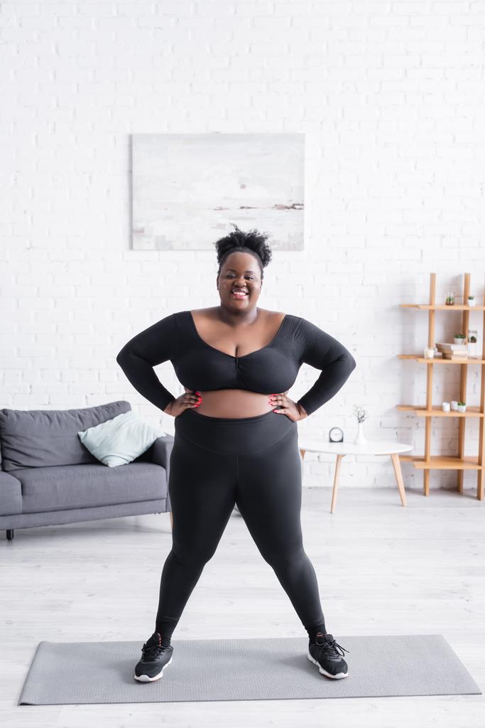 pełna długość zadowolonej afrykańskiej kobiety z nadwagą w odzieży sportowej stojącej z rękami na biodrach w domu - Zdjęcie, obraz
