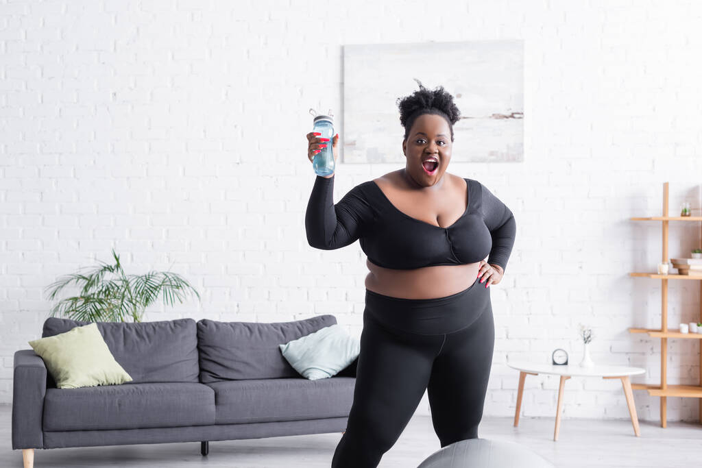 stupito africano americano plus size donna in abbigliamento sportivo in possesso di bottiglia sportiva a casa - Foto, immagini