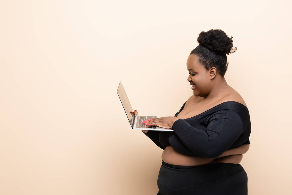 vista lateral da feliz afro-americana plus size mulher segurando laptop isolado no bege  - Foto, Imagem
