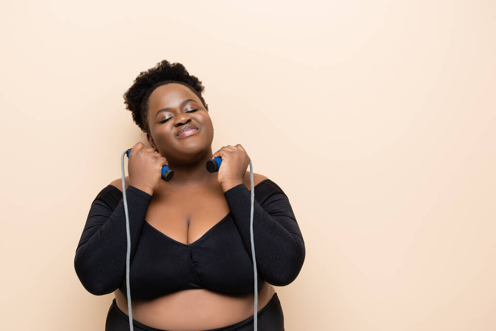 pleased african american plus size woman in sportswear holding skipping rope isolated on beige  - Photo, Image
