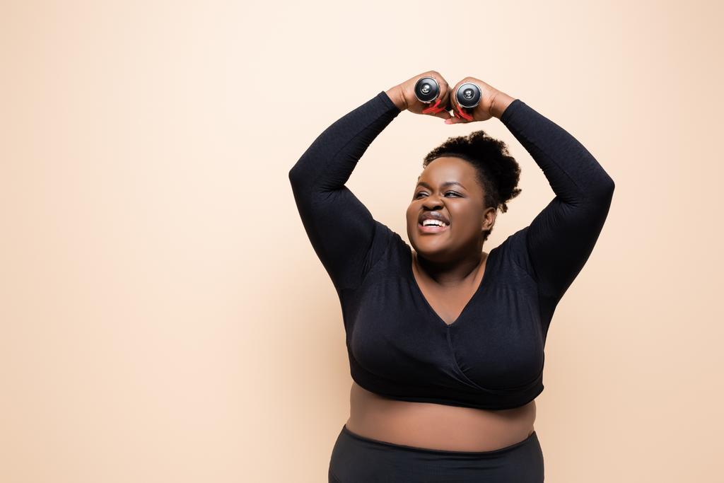 happy african american plus size woman in sportswear holding dumbbells above head isolated on beige  - Photo, Image