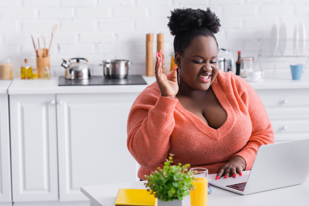heureux afro-américain plus la taille pigiste avec la main levée à l'aide d'un ordinateur portable à la maison - Photo, image