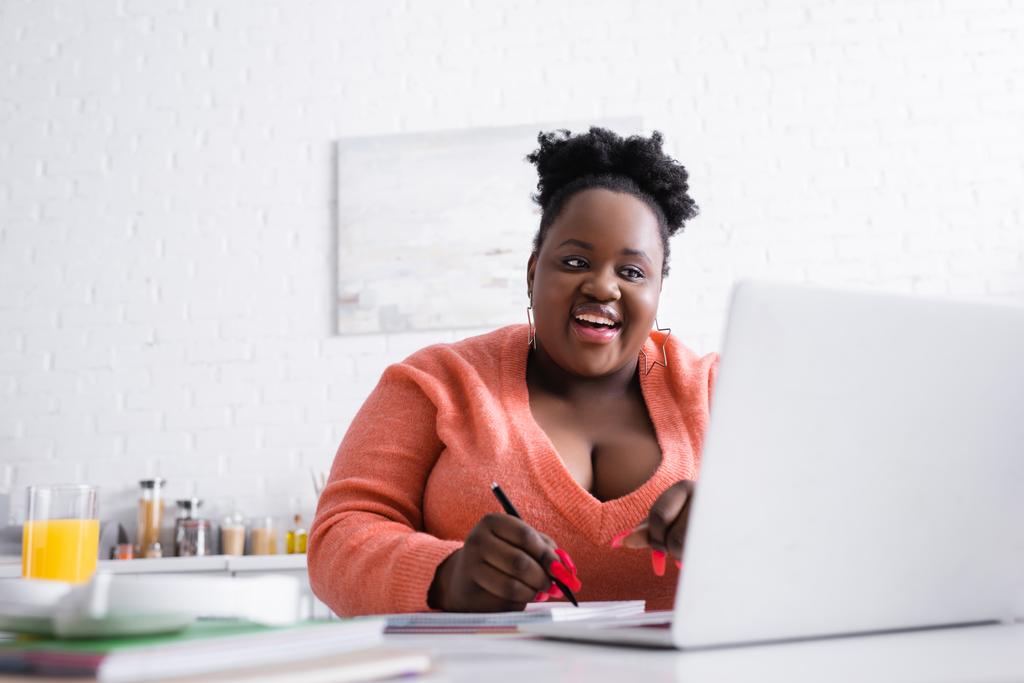веселый африканский американец плюс размер женщина держит ручку и глядя на ноутбук - Фото, изображение