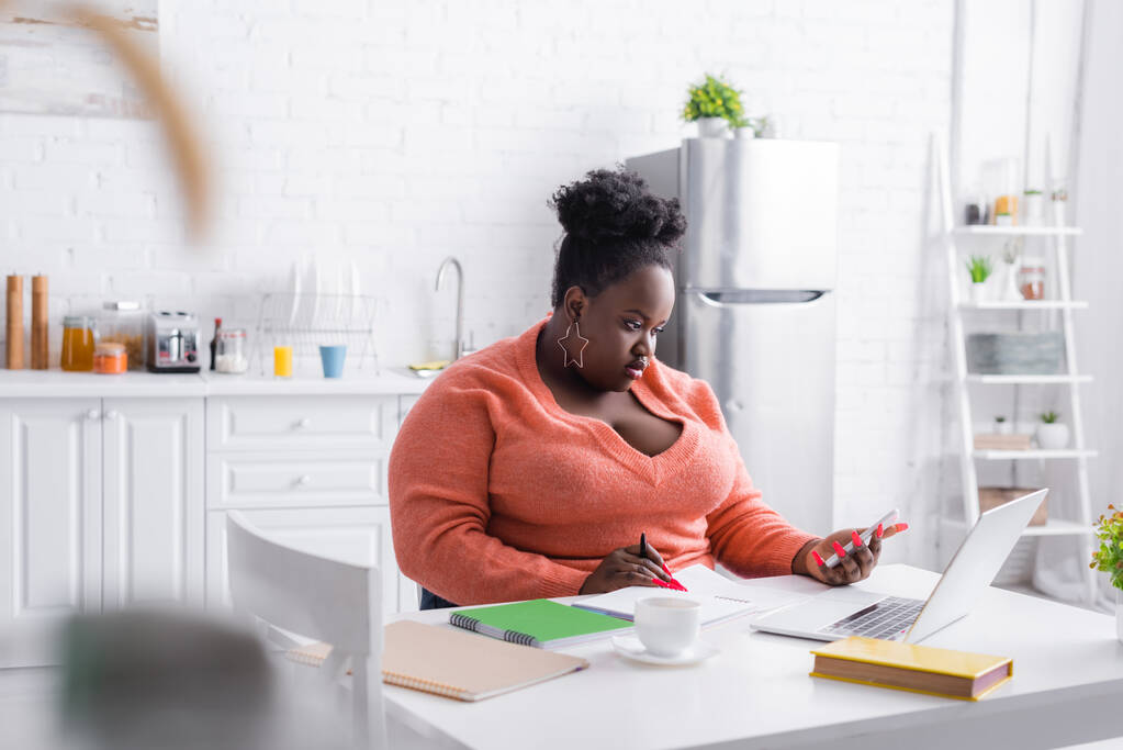 Afroamerykanka plus size kobieta trzyma smartfona w pobliżu laptopa na stole - Zdjęcie, obraz