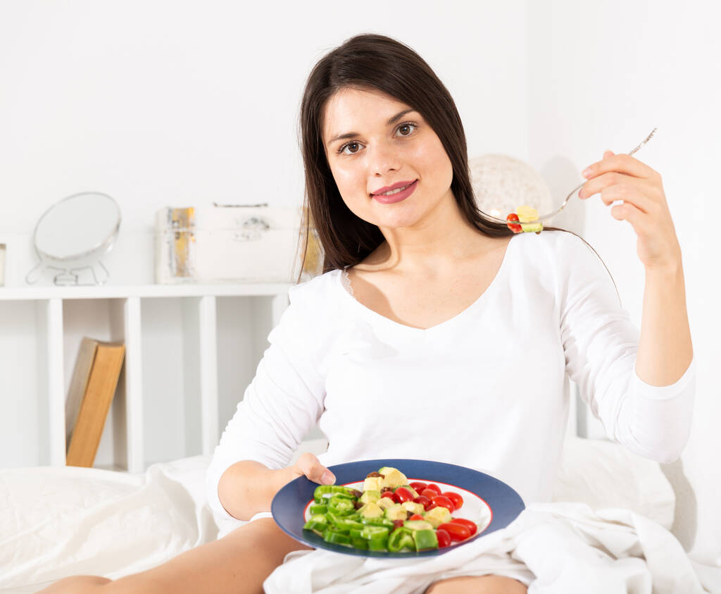 Kaunis nainen valkoisessa mekossa syö kasvissalaattia sängyssä - Valokuva, kuva