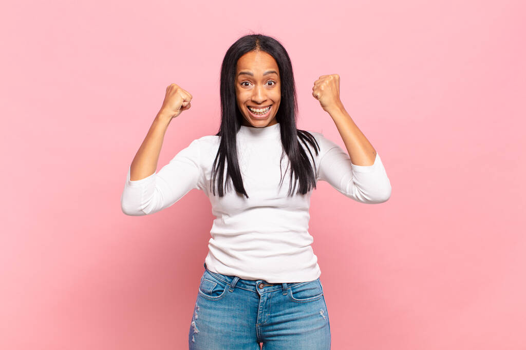 молодая черная женщина чувствует себя счастливой, удивленной и гордой, крича и празднуя успех с большой улыбкой - Фото, изображение