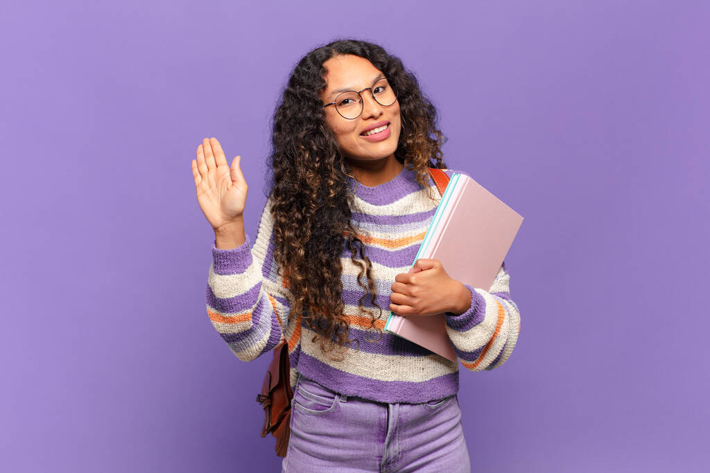young hispanic woman smiling happily and cheerfully, waving hand, welcoming and greeting you, or saying goodbye. student concept - Photo, Image