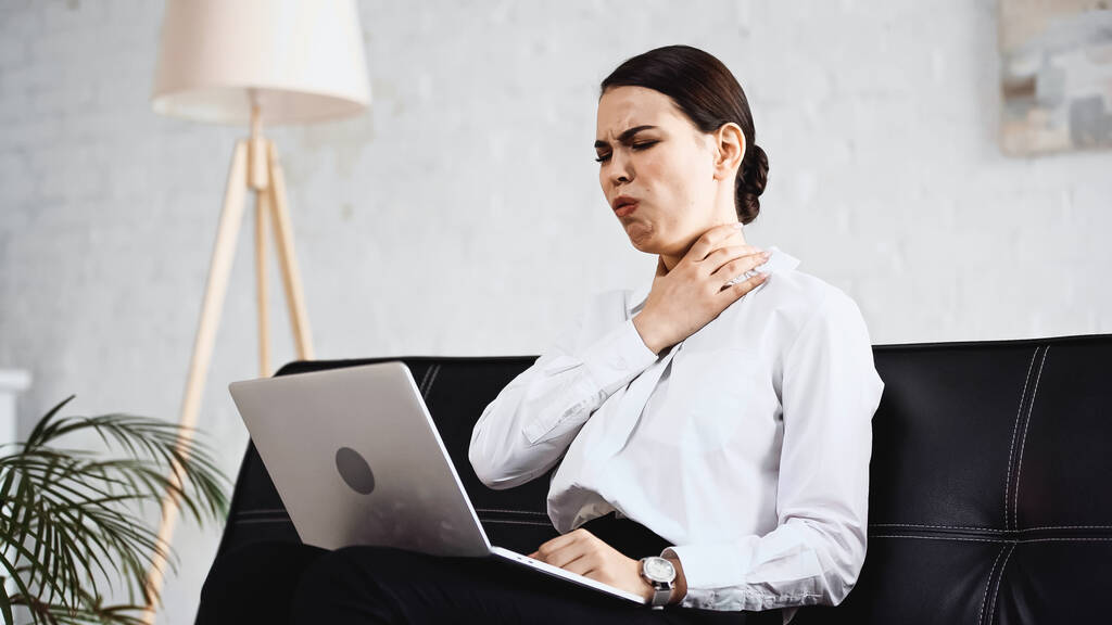 diseased businesswoman touching sore throat while sitting on sofa with laptop - Photo, Image