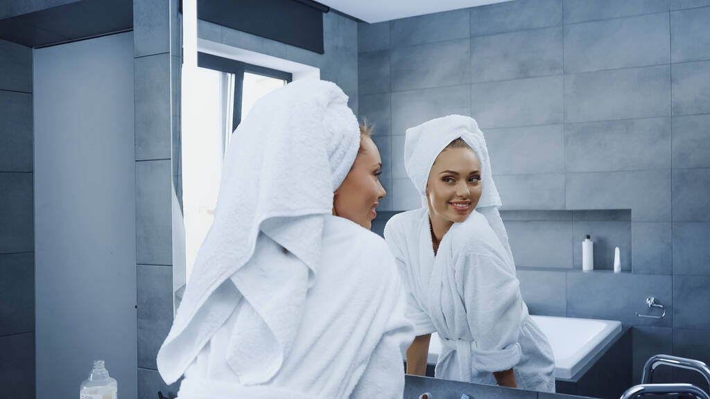 バスルームの鏡を見て頭にタオルを持つ陽気な女性  - 写真・画像