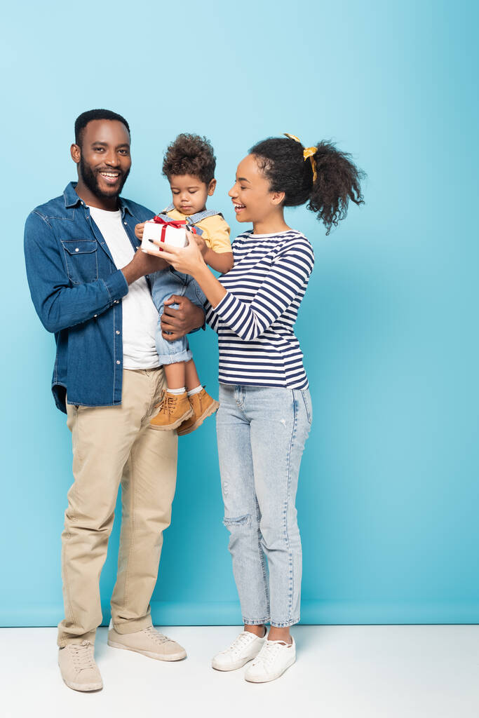 african american man smiling at camera while holding son with gift box near happy wife on blue - Photo, Image