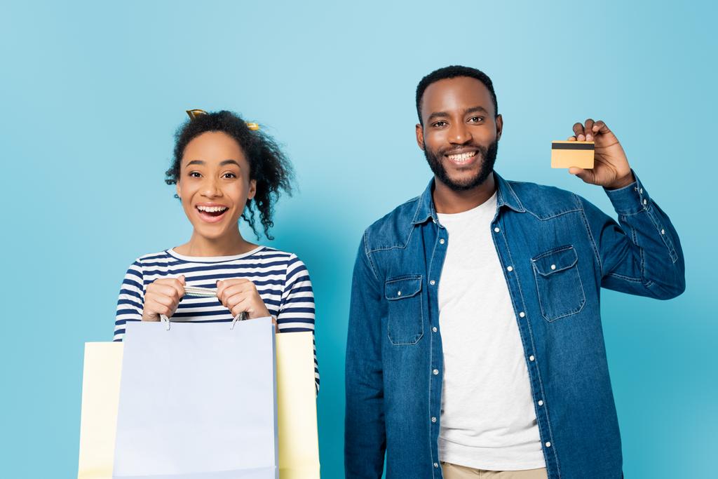 homem americano africano alegre mostrando cartão de crédito perto da esposa animado segurando sacos de compras em azul - Foto, Imagem