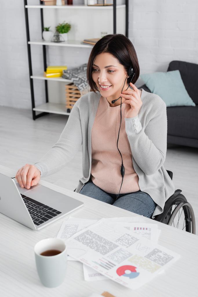 Freelancer deficiente sorrindo no fone de ouvido usando laptop perto de papéis e café em primeiro plano desfocado  - Foto, Imagem