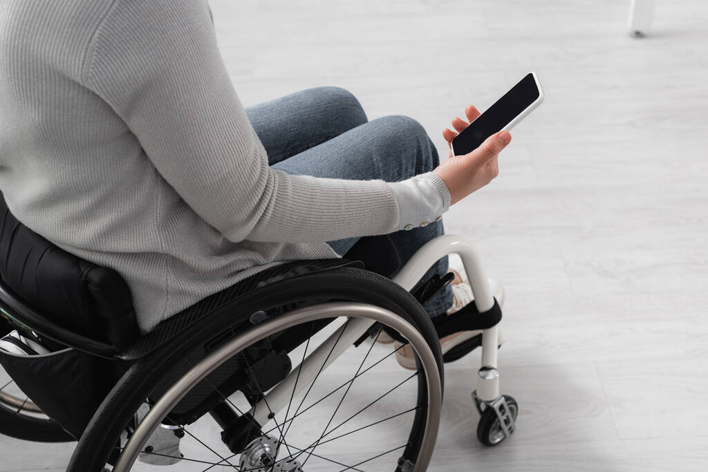 Обрізаний вид жінки з обмеженими можливостями в інвалідному візку, що тримає смартфон з порожнім екраном вдома
  - Фото, зображення