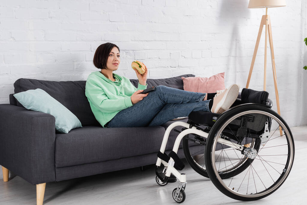 Femme souriante tenant un hamburger tout en regardant un film près d'un fauteuil roulant  - Photo, image