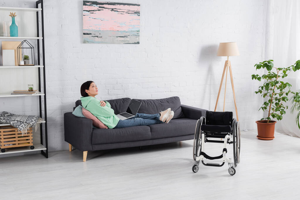 Niepełnosprawny wolny strzelec z laptopem siedzącym na kanapie w pobliżu wózka inwalidzkiego  - Zdjęcie, obraz