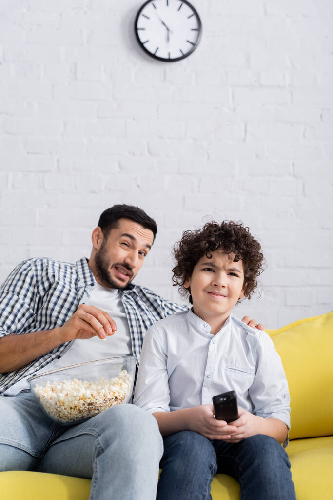 przestraszony arabski człowiek jedzący popcorn podczas oglądania telewizji z synem w domu - Zdjęcie, obraz