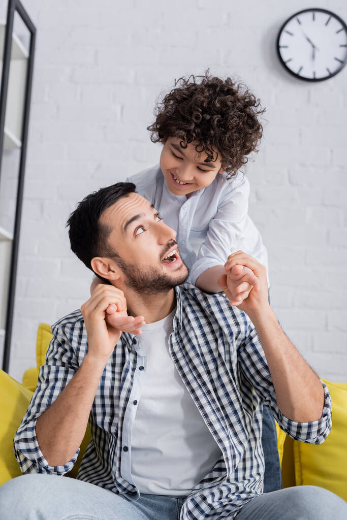 excitado árabe hombre cogido de la mano de feliz hijo mientras se divierten en casa - Foto, Imagen