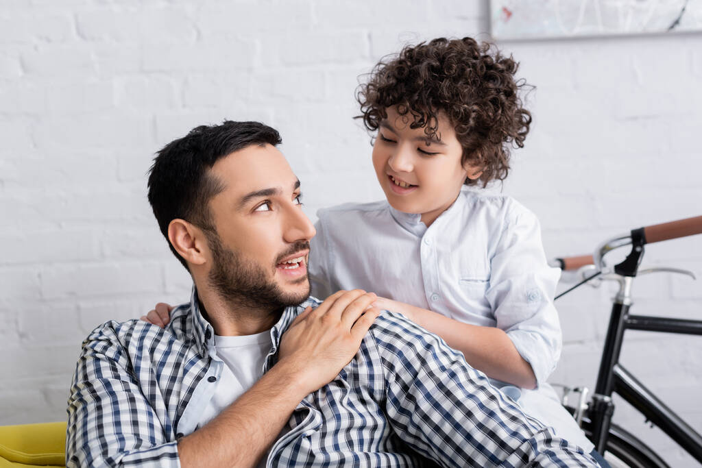 homem árabe feliz tocando a mão do filho sorridente enquanto fala com ele em casa - Foto, Imagem
