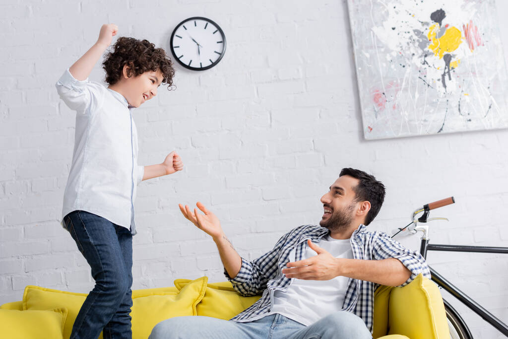homem árabe alegre apontando para o filho animado pulando no sofá com punhos apertados - Foto, Imagem