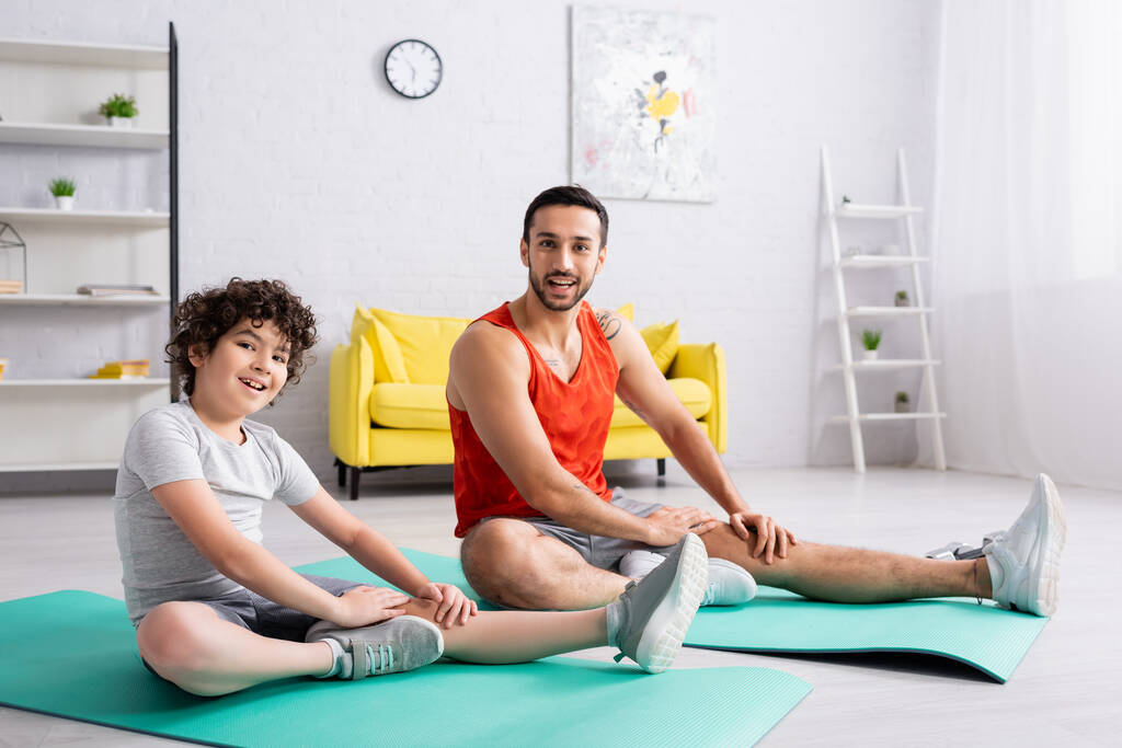 Smiling muslim son and father looking at camera while training on fitness mats  - Photo, Image