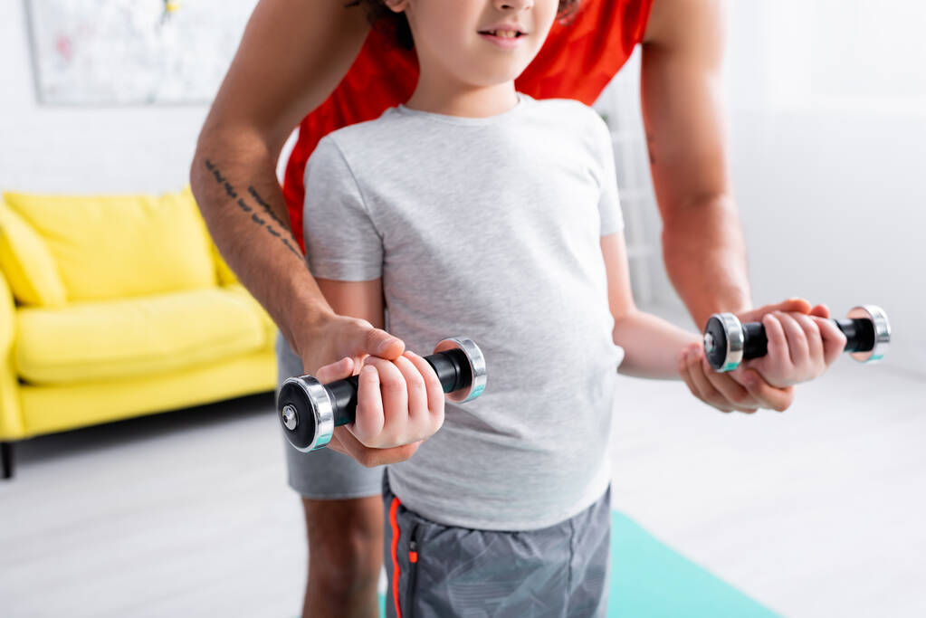 Cropped view of father helping son with dumbbells during work out at home  - Photo, Image