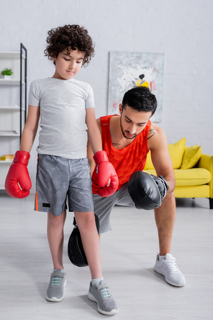 Arabische man in punch mitts onderwijs zoon in bokshandschoenen tijdens het sporten thuis  - Foto, afbeelding