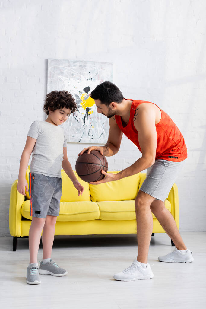 Muslimischer Mann in Sportbekleidung hält Basketball in der Nähe seines Sohnes zu Hause  - Foto, Bild