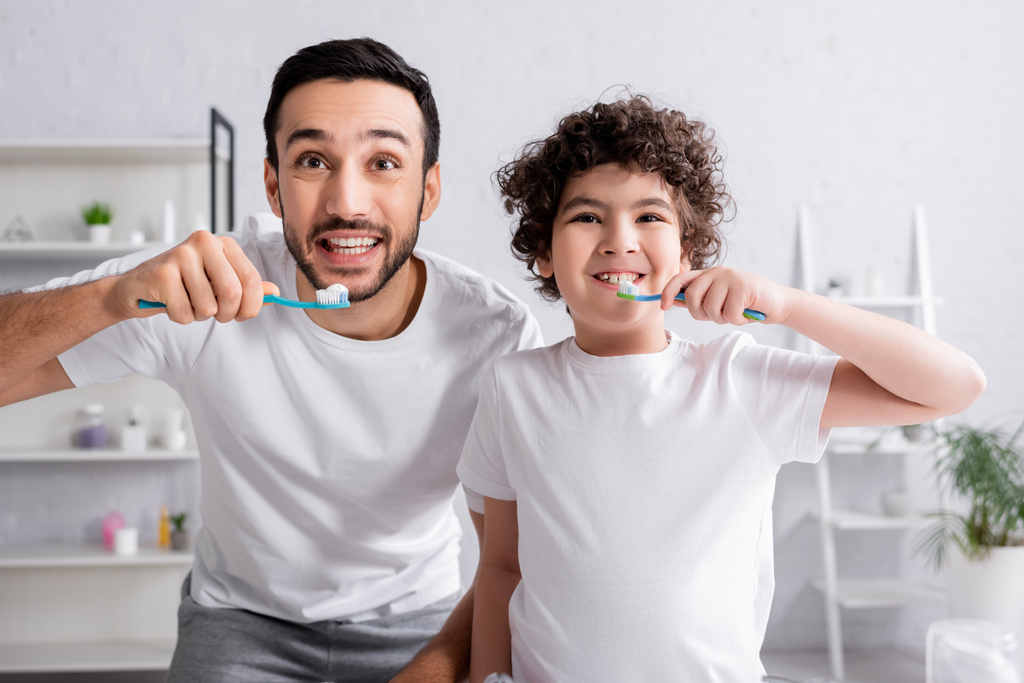 Smiling muslim father and son holding toothbrushes with toothpaste in bathroom  - Photo, Image