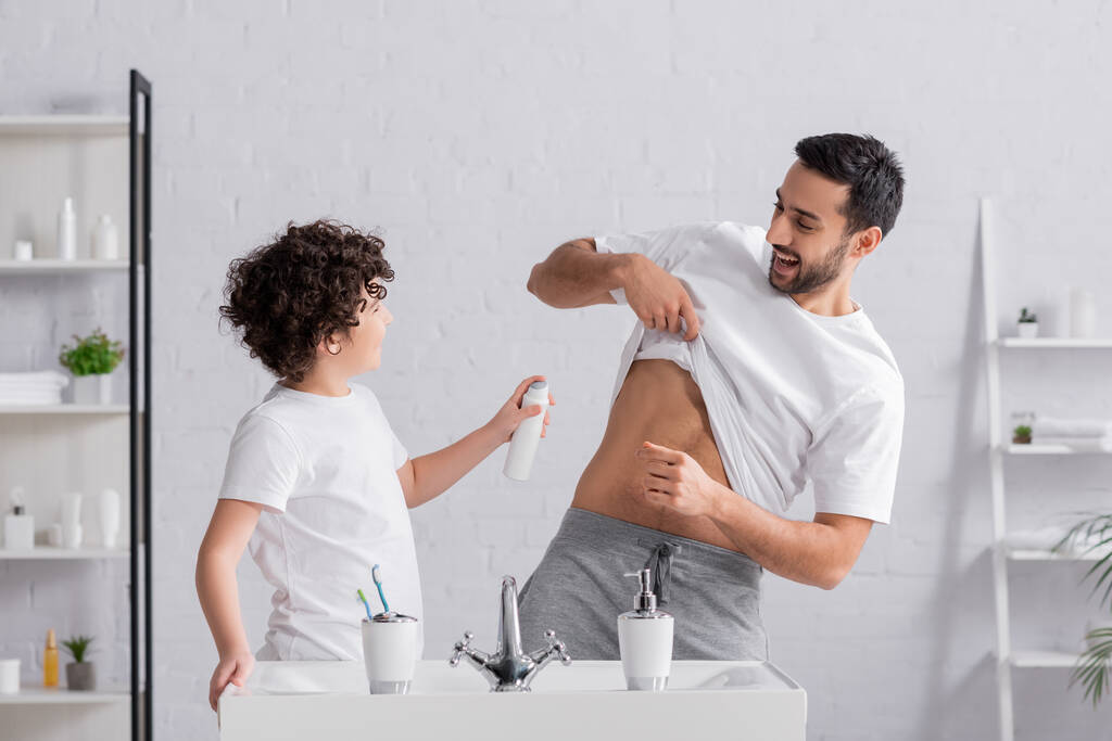 Hombre árabe alegre levantando camiseta cerca de hijo con desodorante - Foto, imagen