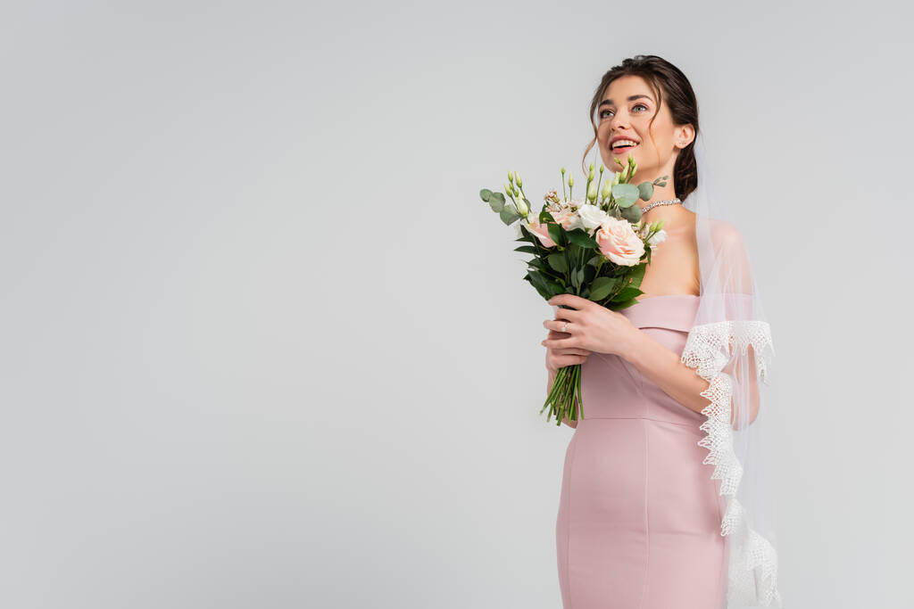 mariée joyeuse regardant loin tout en se tenant debout avec bouquet de mariage isolé sur gris - Photo, image
