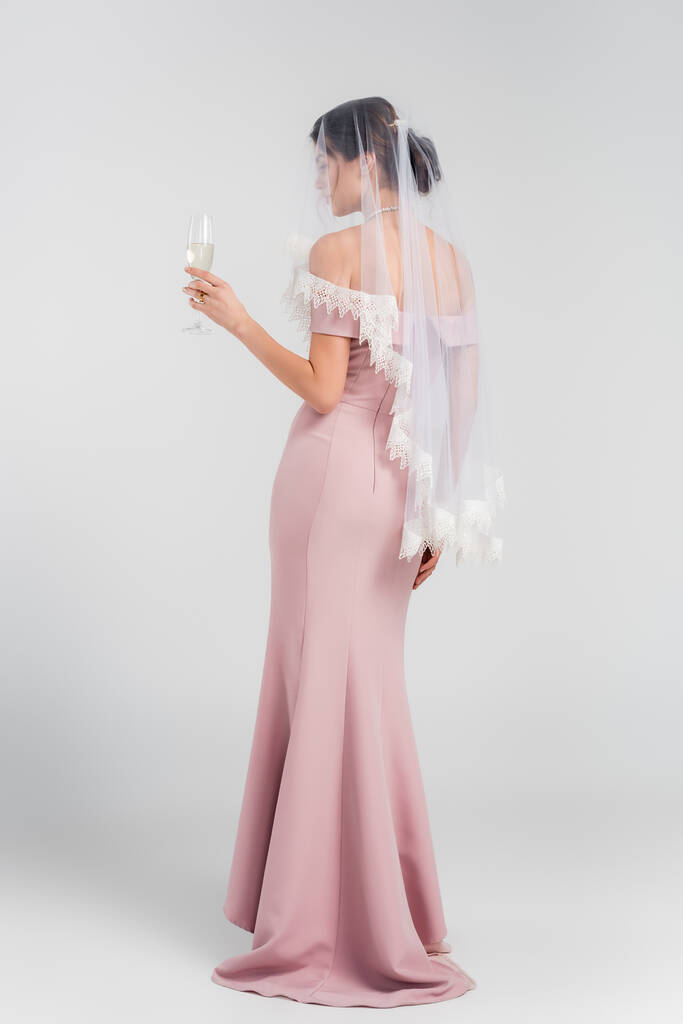 Ganzkörperansicht einer Frau in Brautkleid und Schleier mit einem Glas Champagner auf grau - Foto, Bild