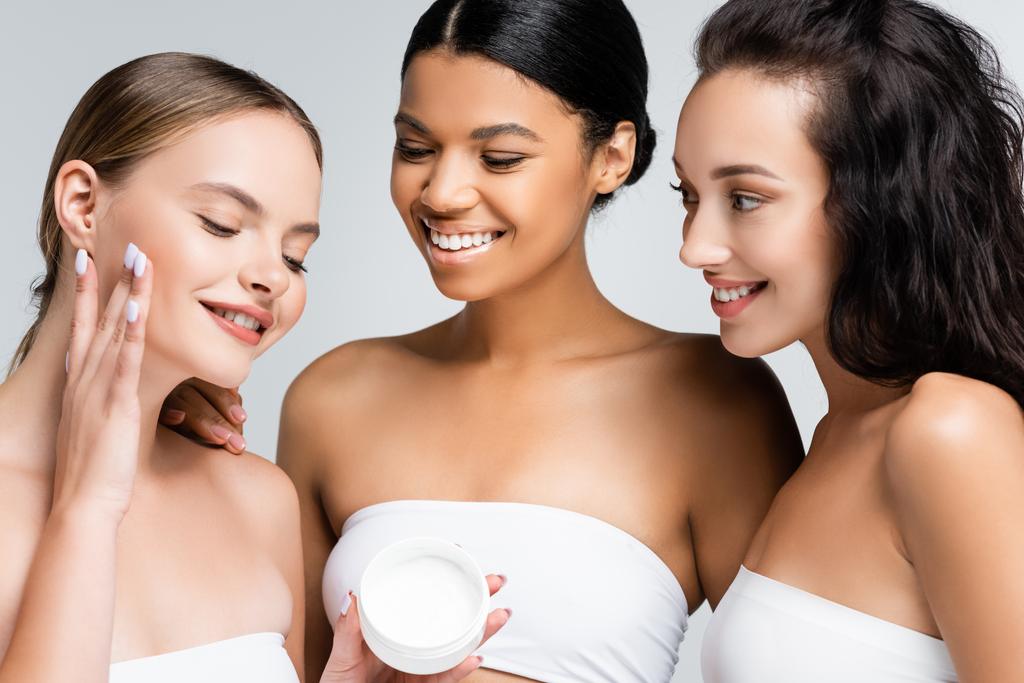 allegre donne multietniche con spalle nude guardando l'amico che applica crema cosmetica isolata sul grigio - Foto, immagini