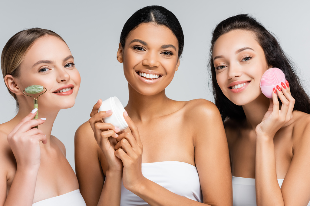 wielonarodowe kobiety uśmiechające się trzymając pojemnik z kremem kosmetycznym, wałkiem nefrytowym i silikonowym środkiem czyszczącym izolowanym na szaro - Zdjęcie, obraz